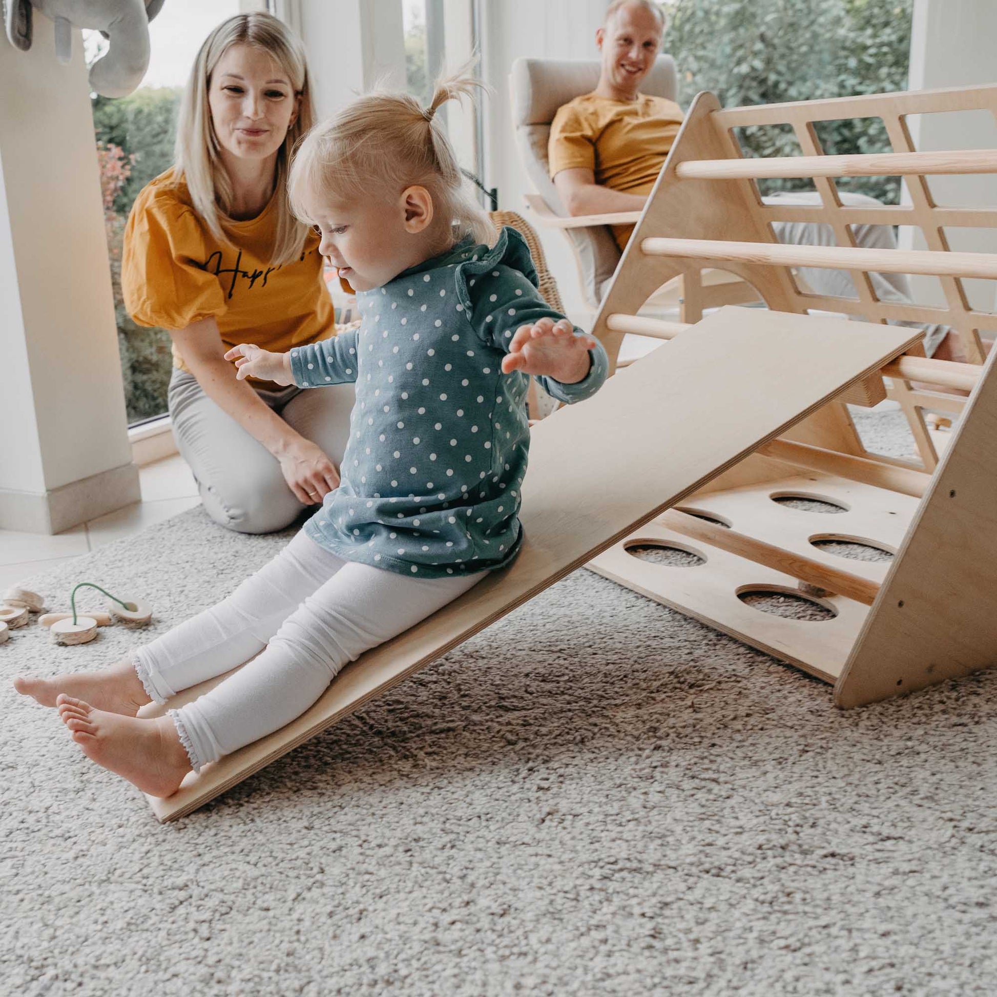 A Montessori-inspired child is playing with a wooden Climbing Triangle + Climbing Arch + a Ramp in a living room.
