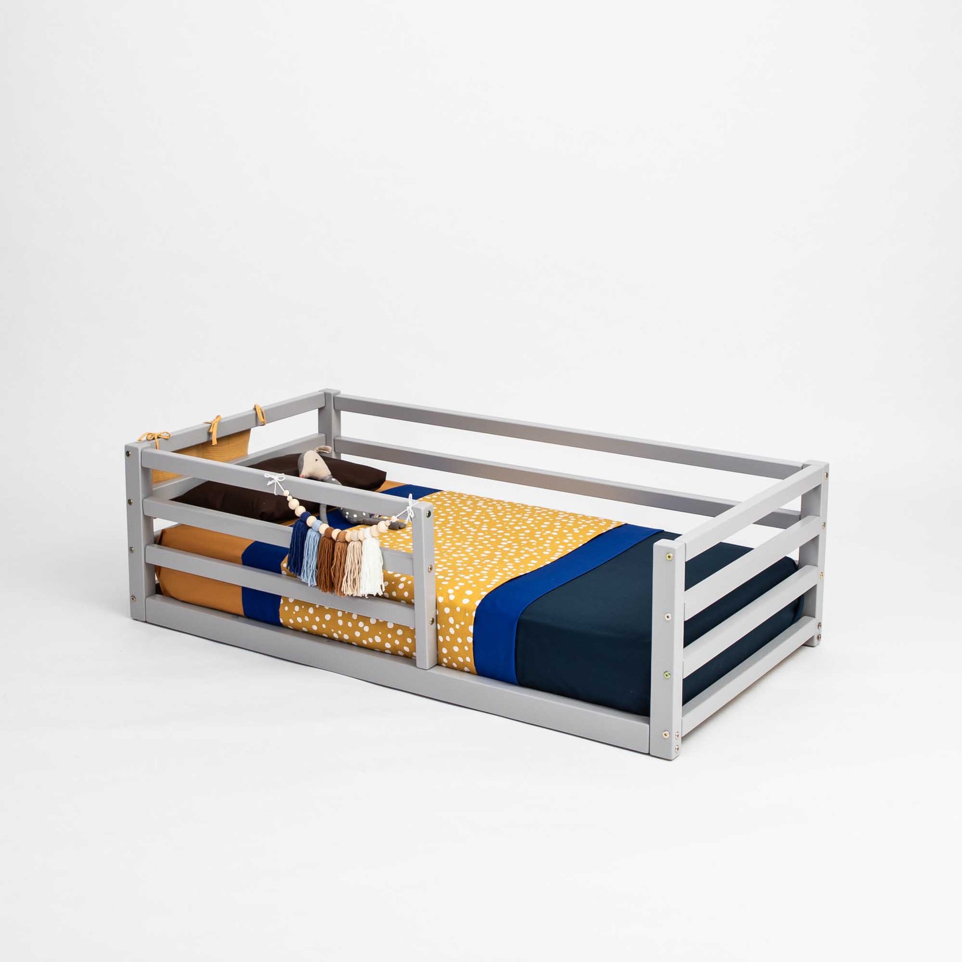 An independent sleeping space for toddlers, the Sweet Home From Wood floor-level kids' bed with a horizontal rail fence features a cozy yellow and blue blanket. It serves as a perfect transitional piece for little ones.