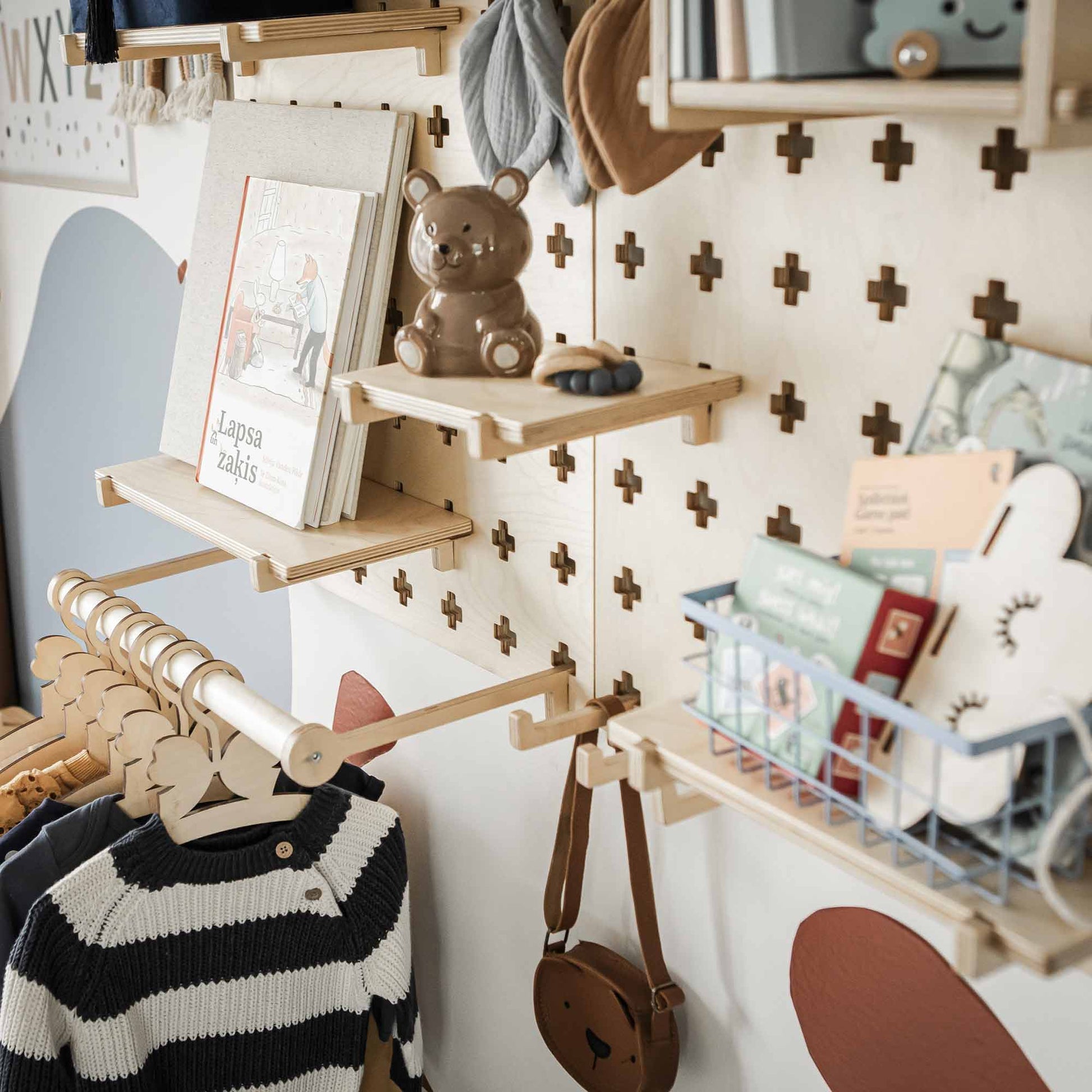 A children's room display features a Large Pegboard Shelf with Clothes Hanger, showcasing books, a teddy bear, clothes on the hanging rod, and other small items. The wall behind sports a cross pattern decorated with toys. This setup serves as an efficient organizer and offers customizable layouts for any space.