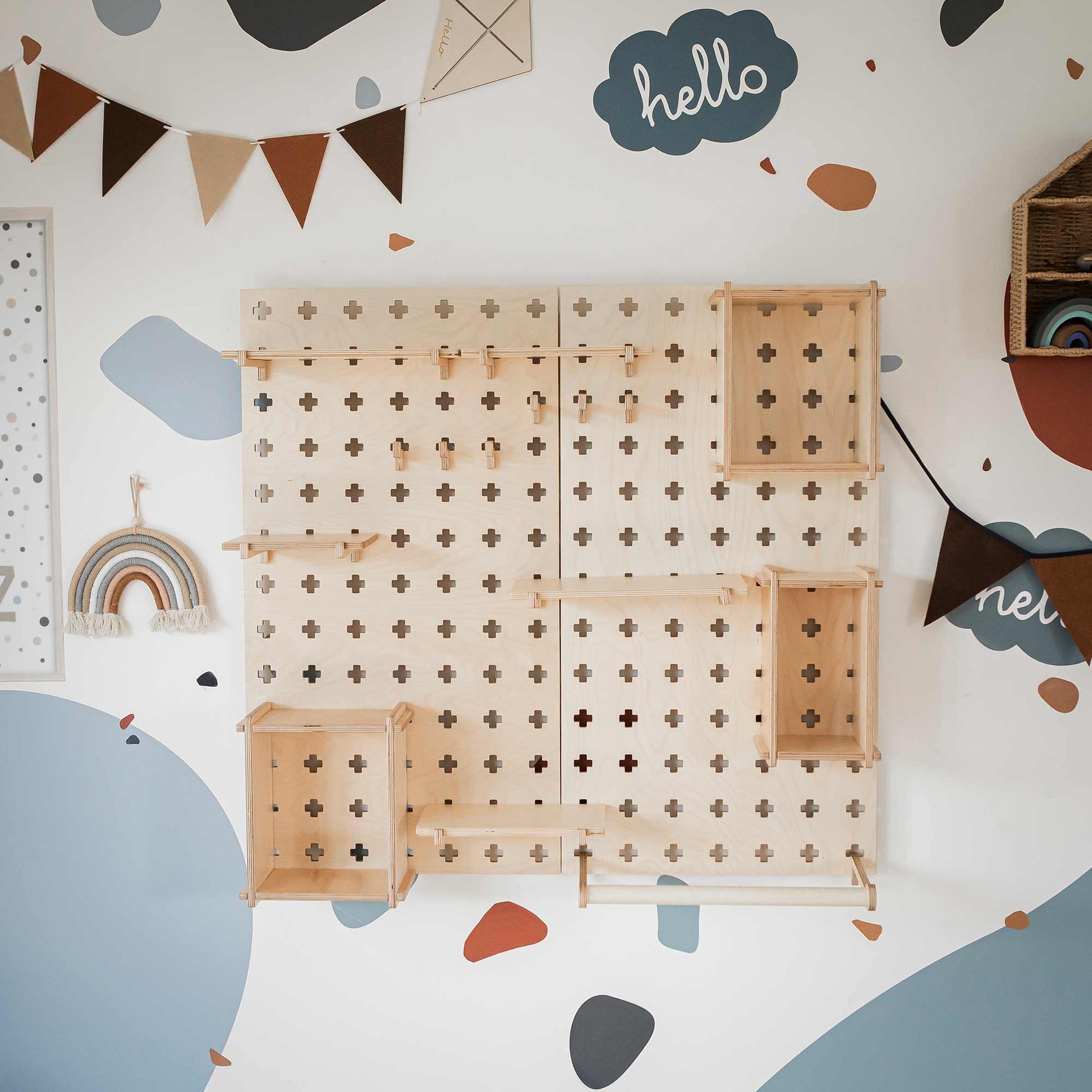 The Large Pegboard Shelf with Clothing Rack, a versatile wall-mounted wooden pegboard, offers customizable layouts with an array of small shelves and pegs. It is displayed against a white wall adorned with bunting, geometric shapes, and a blue "hello" sign.