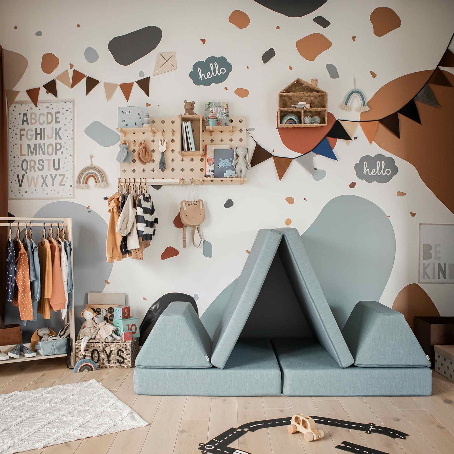 A children's playroom featuring an activity play couch set, colorful abstract wall decals, an alphabet chart, a clothes rack, and a triangular foam play structure creates a vibrant environment. A cozy play tent enhances the playful atmosphere while various toys and decorations are neatly arranged around the room—an ideal Montessori gift for imaginative young minds.