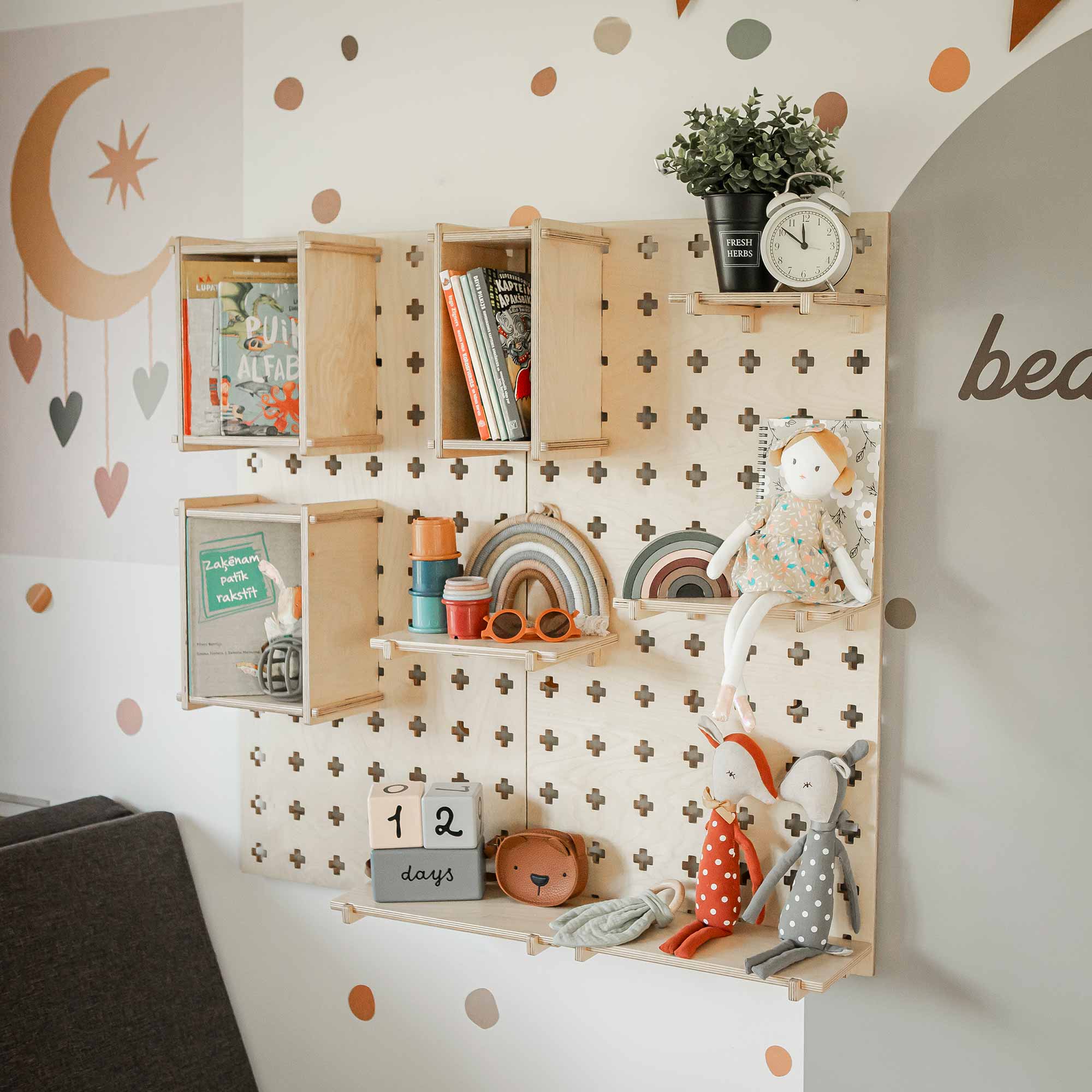 Wall-mounted Large Pegboard Floating Display Shelf holding a variety of children's books, toys, and decor items, including a rainbow, clock, and dolls, create an ideal storage solution in a pastel-colored nursery with polka dot and moon-themed wall art.