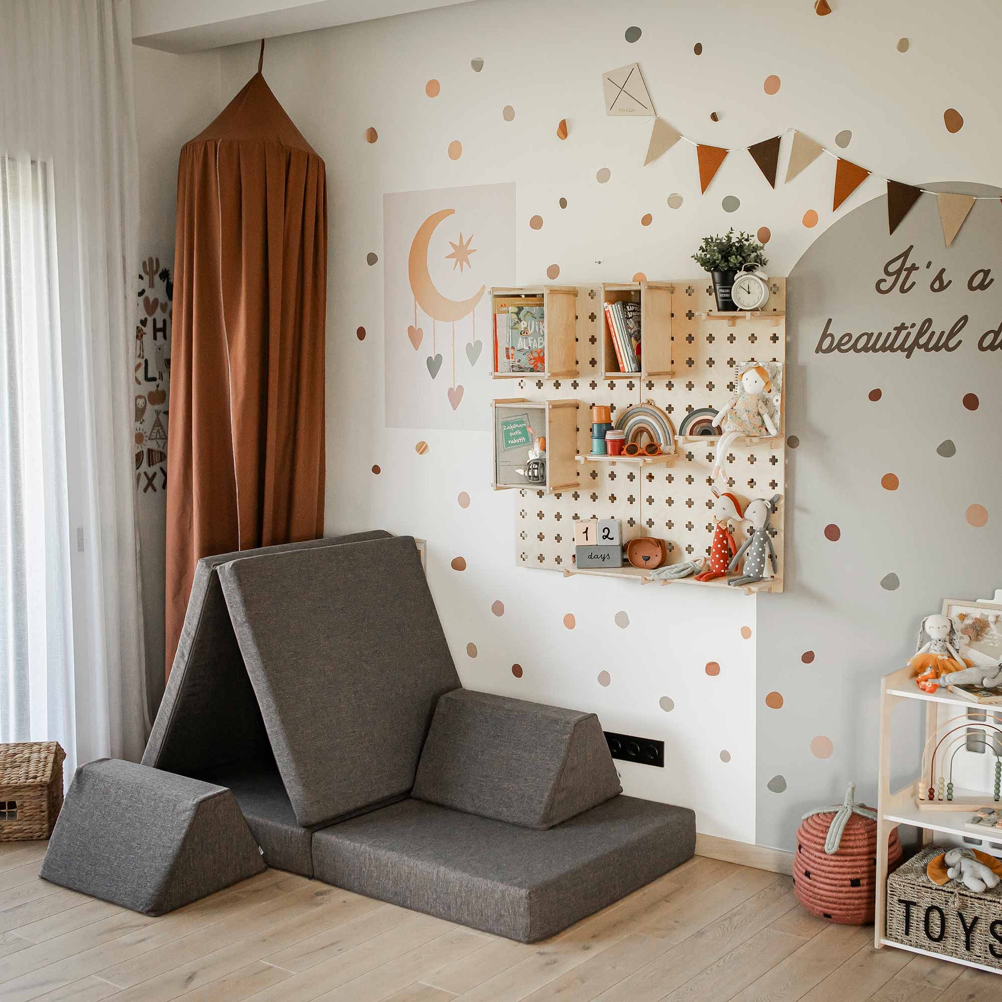 A children's playroom with polka dot walls, a Large Pegboard Floating Display Shelf for organized storage holding toys and decorations, a beige canopy, and folded grey foam mats. A garland and framed prints adorn the walls. A basket sits on the floor.