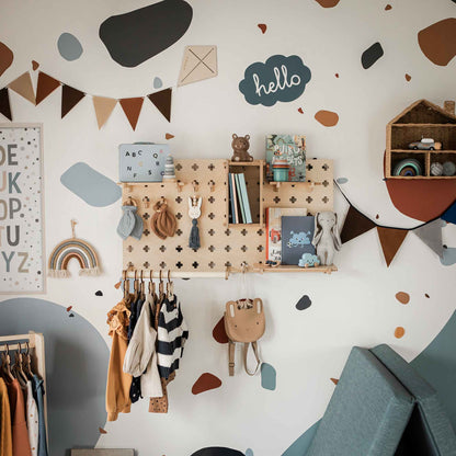 A children's room wall is beautifully organized with the Pegboard with Clothes Clothing Rack, where hanging clothes, toys, and books find their place on wooden pegs and adjustable shelves. The wall boasts vibrant abstract shapes, a small banner, and a "hello" sign. This setup ensures easy maintenance while keeping everything tidy and within reach.