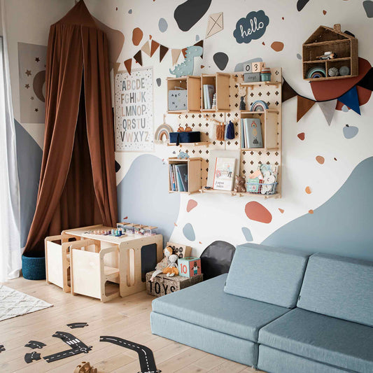 A children's playroom with a blue sofa features a Montessori weaning table and 2 chair set, perfect for fostering independence. Wall-mounted shelves hold various toys, aiding in fine motor skill development, while a colorful patterned wall and a canopy tent in one corner add charm.