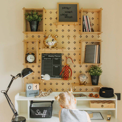 A child sits at a desk with organized shelves, a weekly planner, a clock, books, and decor items including a piggy bank and plants. There's a lamp, a "Grade 1" sign, and a bag labeled "Stuff" on the desk. The Large Pegboard Shelf with Floating Shelves provides customizable layouts for premium storage solutions.