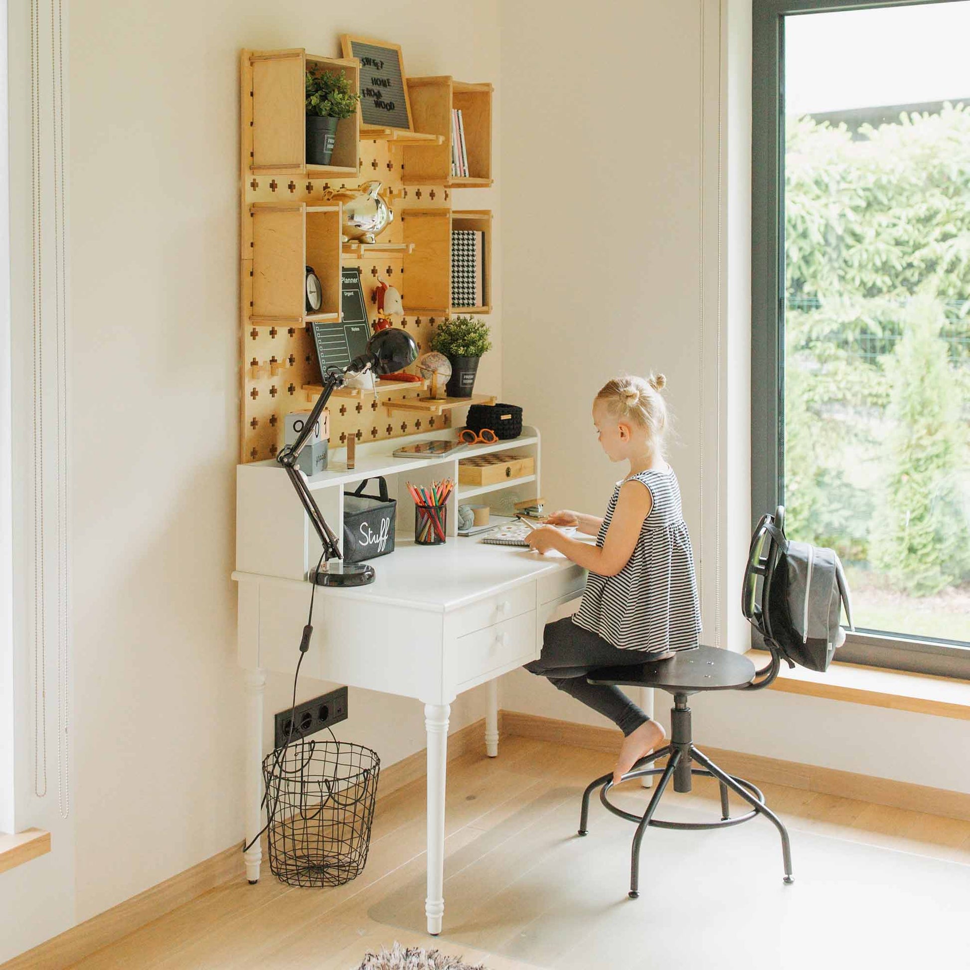 A child sits at a Large Pegboard Shelf with Floating Shelves desk, using a computer in a well-lit room with large windows overlooking greenery. The organized shelves provide customizable layouts and premium storage solutions.
