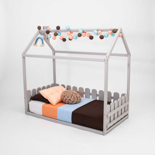 The Floor house floor bed with 3-sided picket fence rails features a charming house-shaped frame, adorned with multicolored string lights and throw pillows on a white background—making it an ideal whimsical toddler bed.