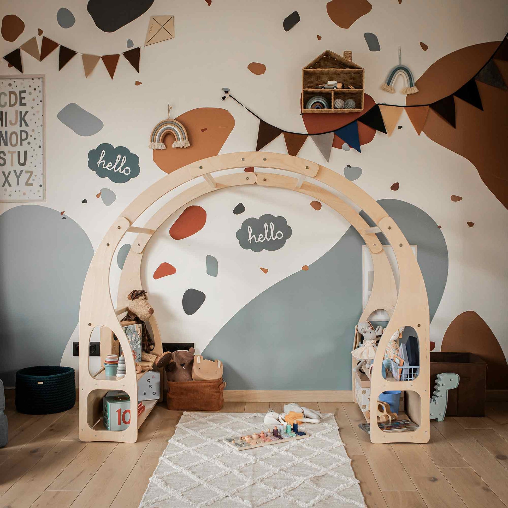 Children's playroom featuring a wooden play stand for toddlers, multi-functional Montessori toys, a rug, alphabet wall art, and bunting decorations. The walls are painted with colorful abstract shapes and feature a "hello" sign. Designed to foster child development and imaginative play.