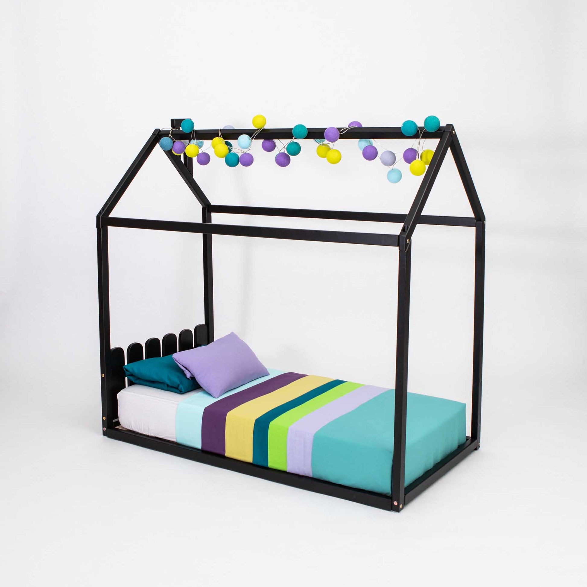 A Kids' house-frame bed with a picket fence headboard with colorful pom poms hanging from it.