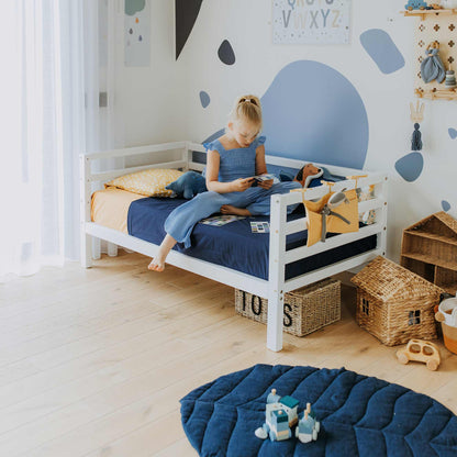 A child sits on a 2-in-1 transformable kids' bed with a 3-sided horizontal rail and solid wood legs in a brightly lit room, reading a book. The room has a blue and white color scheme, decorated with toys, wicker baskets, and a blue cushioned mat on the wooden floor.