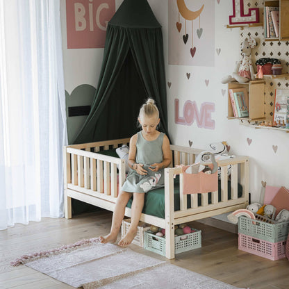 A young girl sits on a sturdy "Toddler Bed on Legs with a Fence" in a decorated room adorned with wall art and toys. The space, designed to encourage independent sleeping, features a cozy canopy, floating shelves, and storage bins filled with playthings.