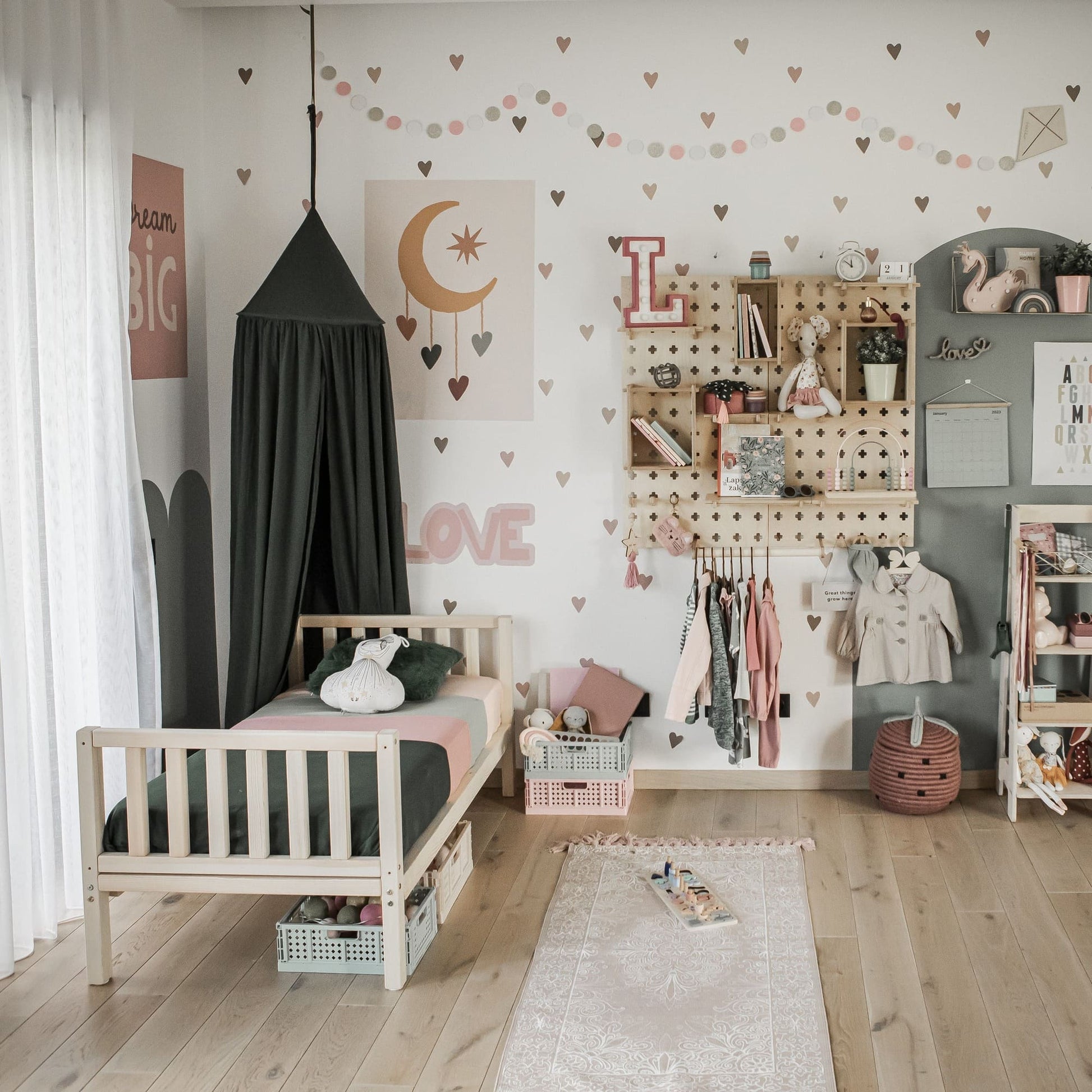 Children's bedroom with a small Montessori-inspired raised kids' bed featuring a vertical rail headboard and footboard. The room includes pink and white decorations, a dark canopy, hanging clothes, wall-mounted storage, and a soft pink rug on the solid wood floor.