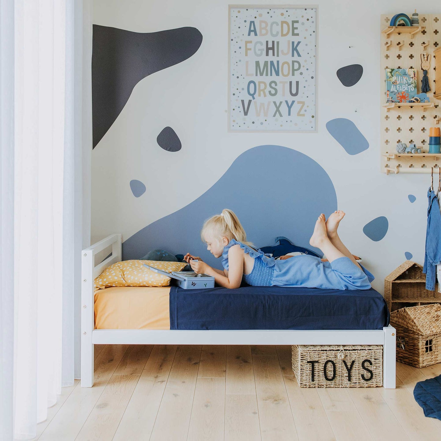 A child lies on their stomach on a Kids' bed on legs with a horizontal rail headboard in a neatly organized room, reading a book. The wall behind them is decorated with abstract shapes and an alphabet poster. A box labeled "toys" is on the floor, beside the bed's solid pine wood sturdy frame.