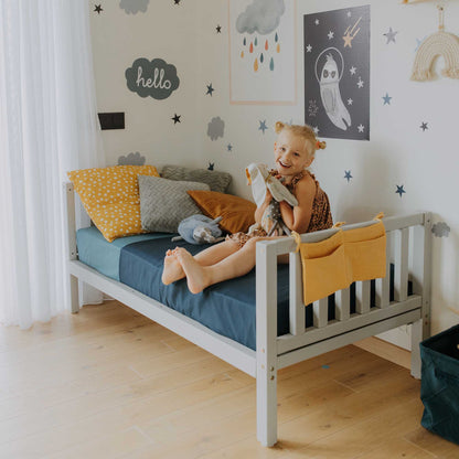 A little girl sitting on a Sweet Home From Wood raised kids' bed on legs with a headboard and footboard in a Montessori-inspired child's room.