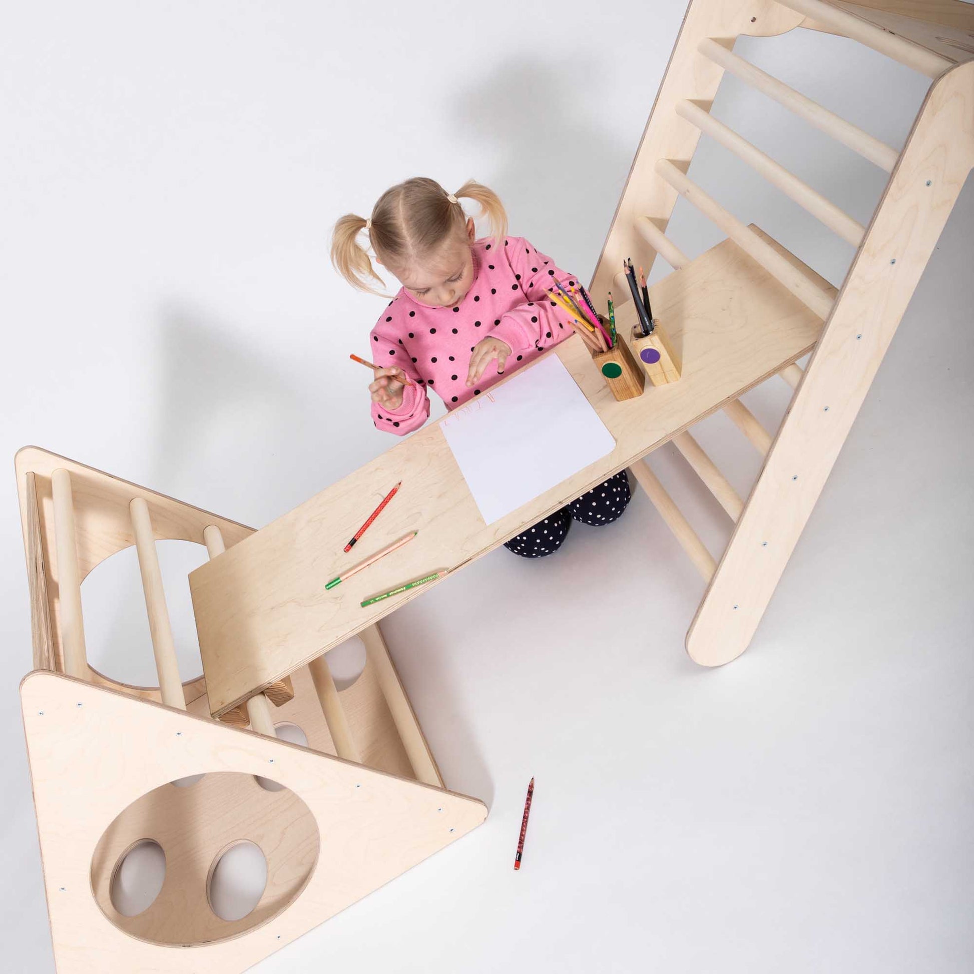 A girl creatively using a climbing triangle ramp as a table for drawing, showcasing the versatility of the structure for alternative activities.
