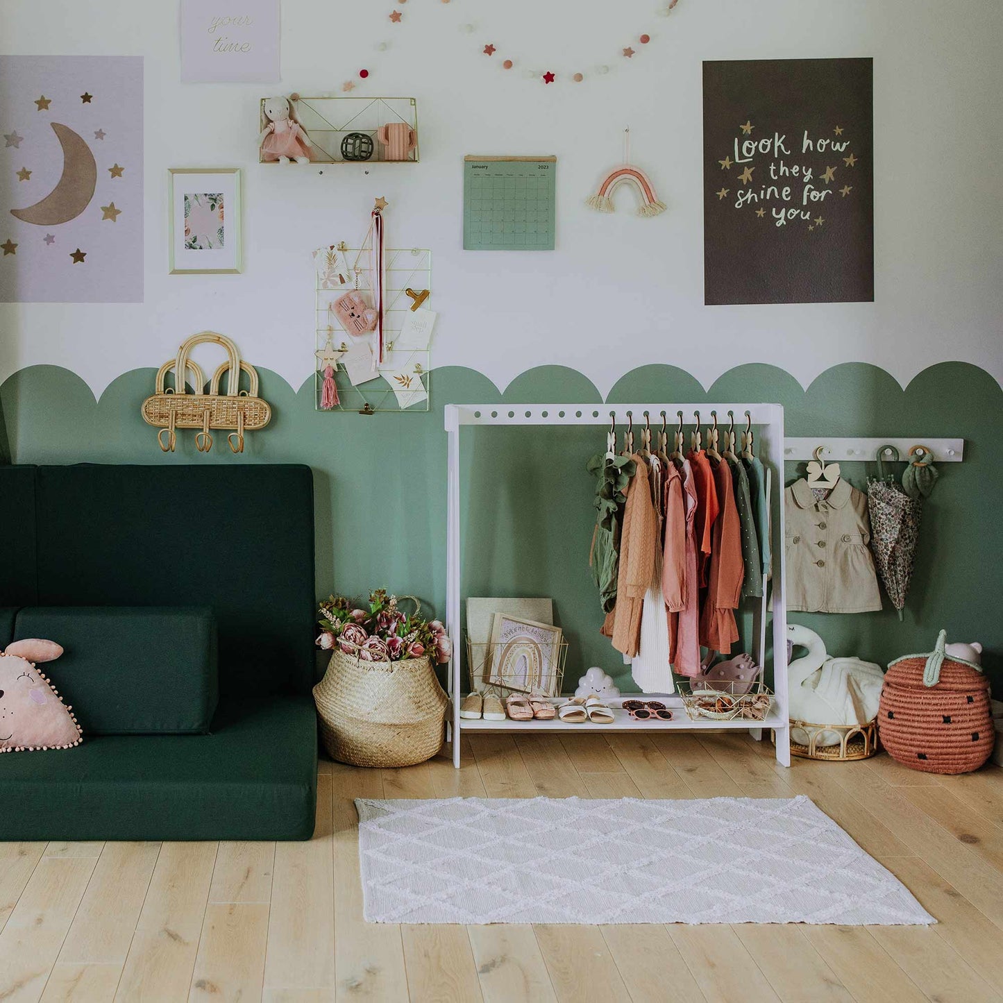 A children's playroom with a green couch, a Montessori clothing rack holding colorful clothes, wall decorations including stars and quotes, and baskets on the floor. A white rug is in front of the rack.