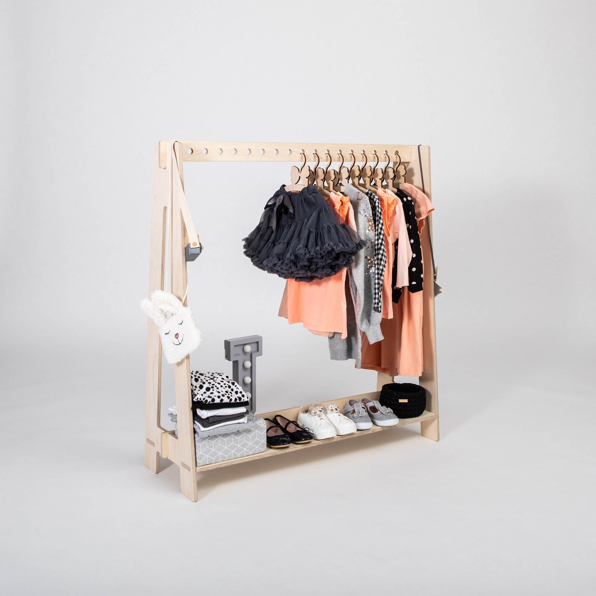 The Montessori clothing rack is a compact, wooden piece that beautifully displays various children's clothes such as dresses, shirts, and jackets. Its bottom shelf is designed to hold shoes and storage boxes for efficient clothing organization.