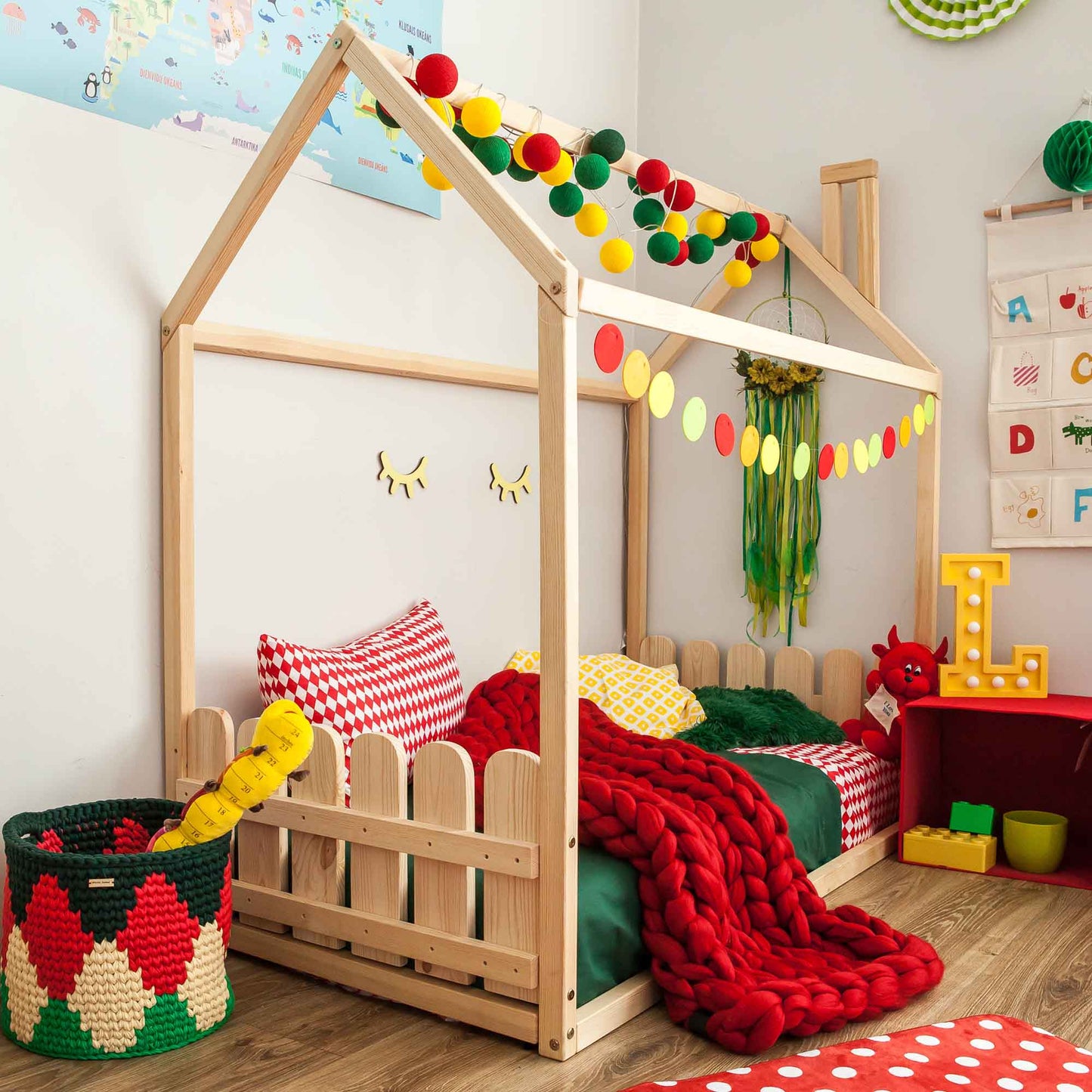 A children's room featuring a Montessori house-frame bed with a picket fence headboard and footboard, perfect for young enthusiasts, adorned with red and green decor and colorful garlands. A basket filled with toys, a red table, and wall decorations complete the playful setup.