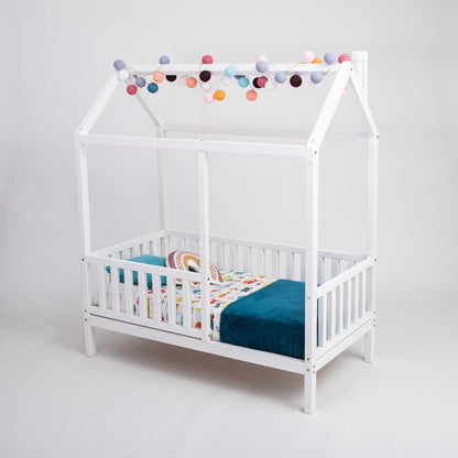 A kids' house bed on legs with a fence with pom poms hanging from the ceiling.