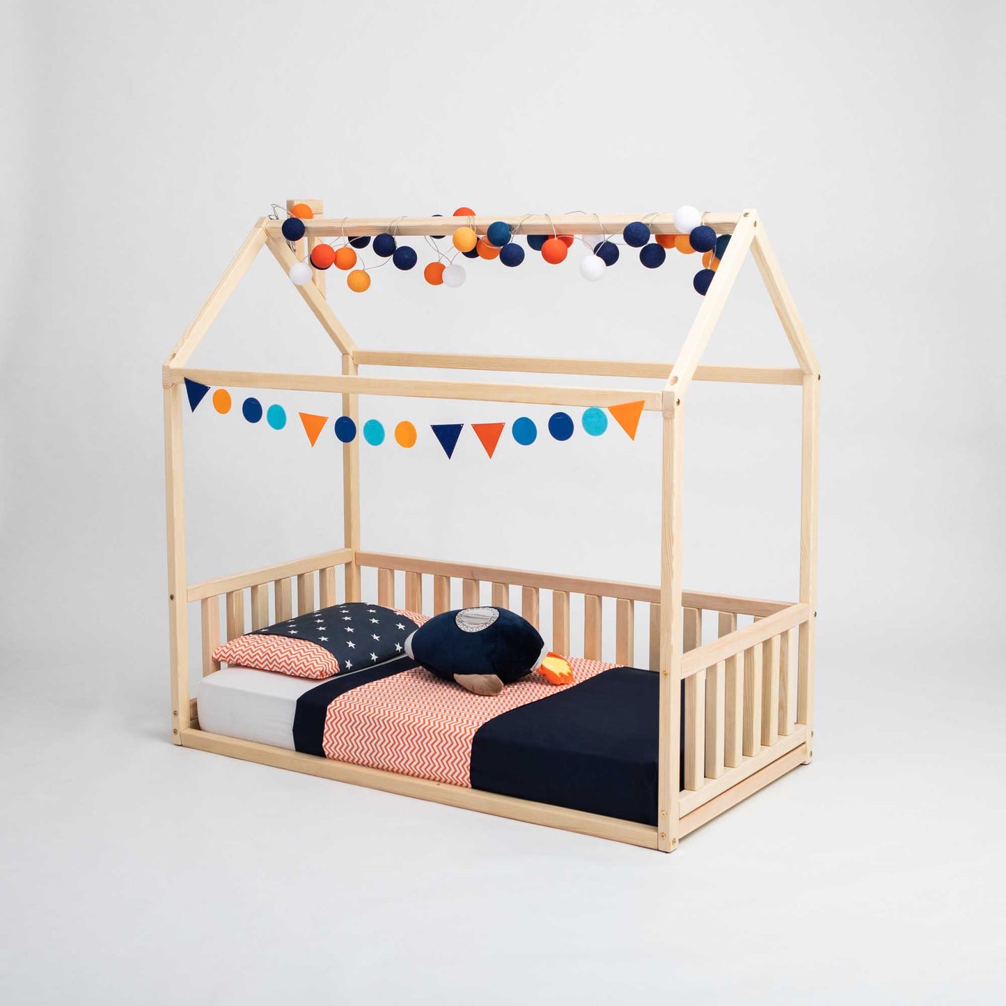 A charming Kids' house-frame bed with 3-sided rails, this wooden toddler bed is shaped like a house and features a house-shaped canopy. It’s beautifully adorned with multicolored bunting, pom-pom garlands, and vibrant blue and orange bedding.