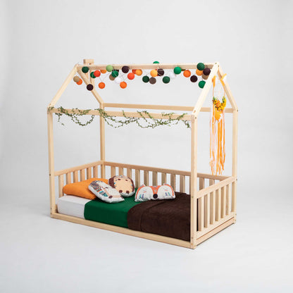 A Sweet Home From Wood Kids' house-frame bed with 3-sided rails, created as a cozy sleep haven, with pom poms hanging from it.