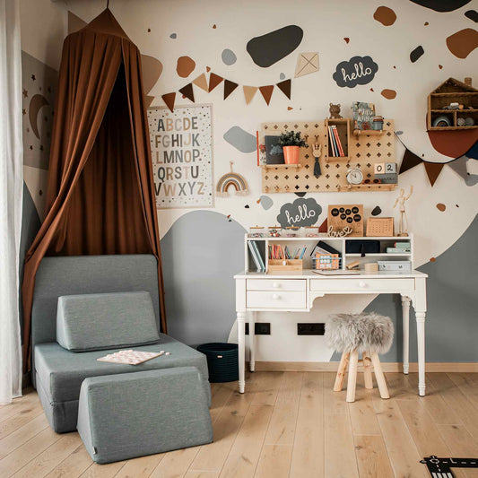 A children's room featuring a grey cushioned chair, a versatile white pedestal desk with a furry stool and storage hutch, wall-mounted decorations, books, and plants. The wall displays a mix of abstract shapes and educational decor elements.
