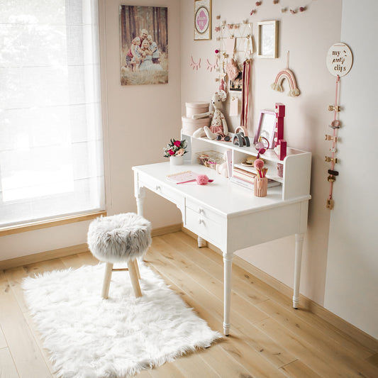 A bright, cozy bedroom corner featuring a versatile white Pedestal desk with decor and a fluffy stool on a white fur rug near a window.