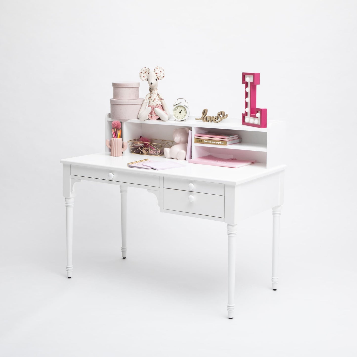 A tidy white versatile Pedestal desk with drawers, accessorized with pink and white objects including books, boxes, a stuffed toy, and a large letter 'L'.