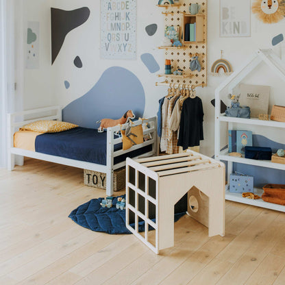 A neatly arranged children's room with a bed, toys, books, a clothing rack, and wall decorations. The decor features a blue and white color scheme with green and yellow accents, including a Montessori table for learning and an activity cube with sensory panels for endless fun.