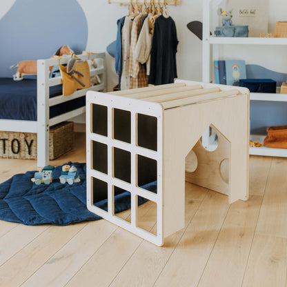 A children's playroom features a wooden play structure as the centerpiece, complemented by a blue quilt, toys, a small bed, and shelves stocked with toddler toys. A clothing rack can be seen in the background. The room also includes an Activity Cube with Sensory Panels and a Montessori table for creative activities, enhancing its educational charm.