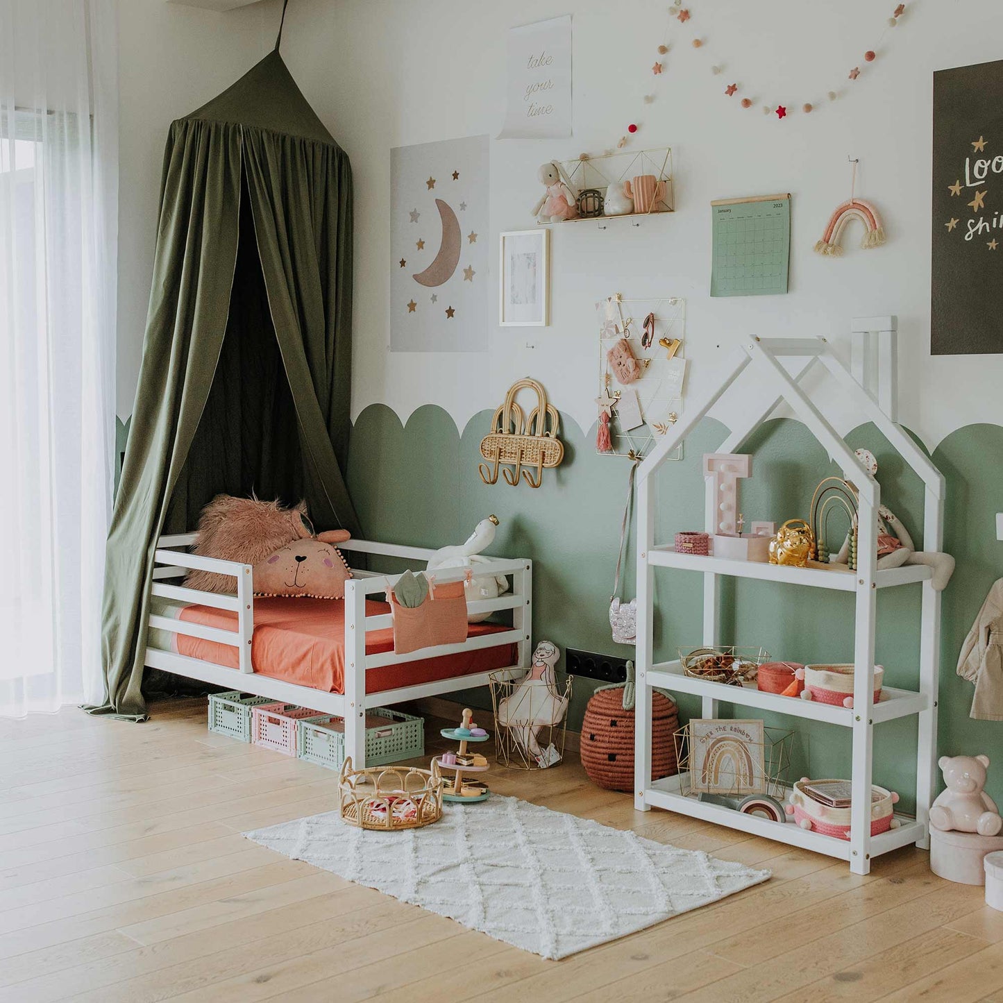 A child's room with a Montessori-style toddler bed, a toy shelf, and wall decorations featuring stars, a rainbow, and a moon. A rug with toys is on the wooden floor, and a green canopy is above the Kids' Bed on Legs with Horizontal Rail Fence.