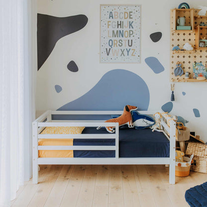 A Kids' bed on legs with a horizontal rail fence is adorned with blue and yellow bedding and is situated against a wall decorated with an alphabet poster and blue abstract shapes. The solid wood frame ensures safety while nearby shelving to the right holds various toys and wicker baskets.