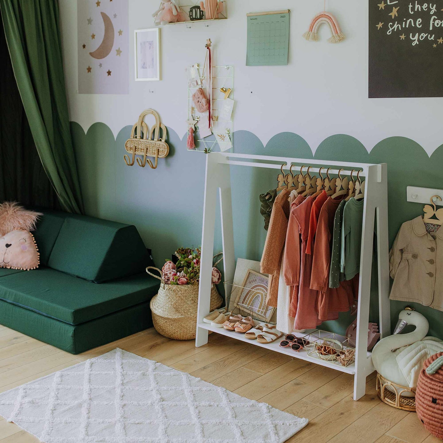 Children's playroom with a dark green couch, a small white rug, and a Kids' clothing rack in A-frame style holding children's clothes. The walls are decorated with various pictures and toys.