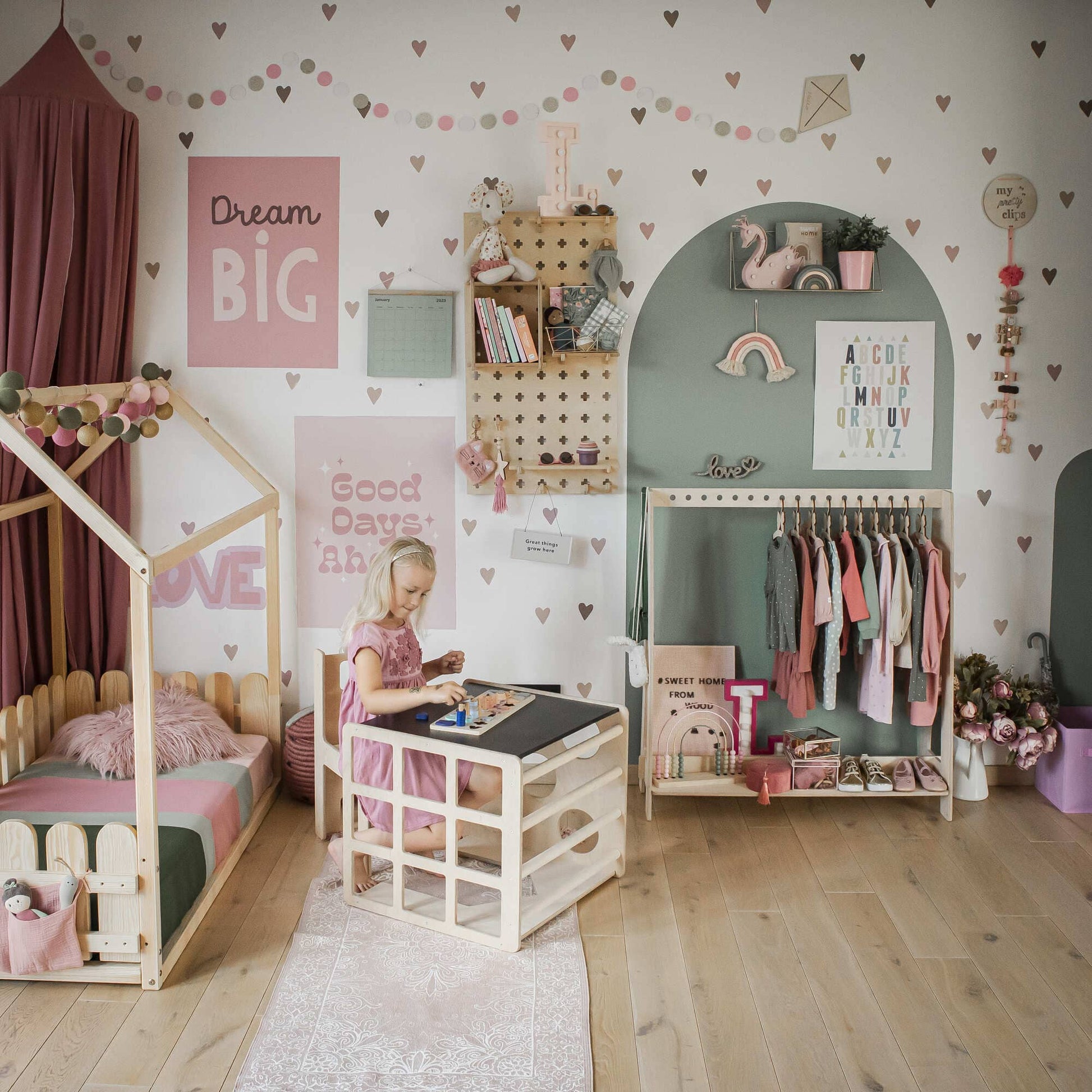 A young toddler engages with toys at a 2-in-1 table and chair set or activity cube in a pastel-themed bedroom, adorned with wall art, a wooden bed, and a clothing rack displaying pink and neutral-colored clothes.