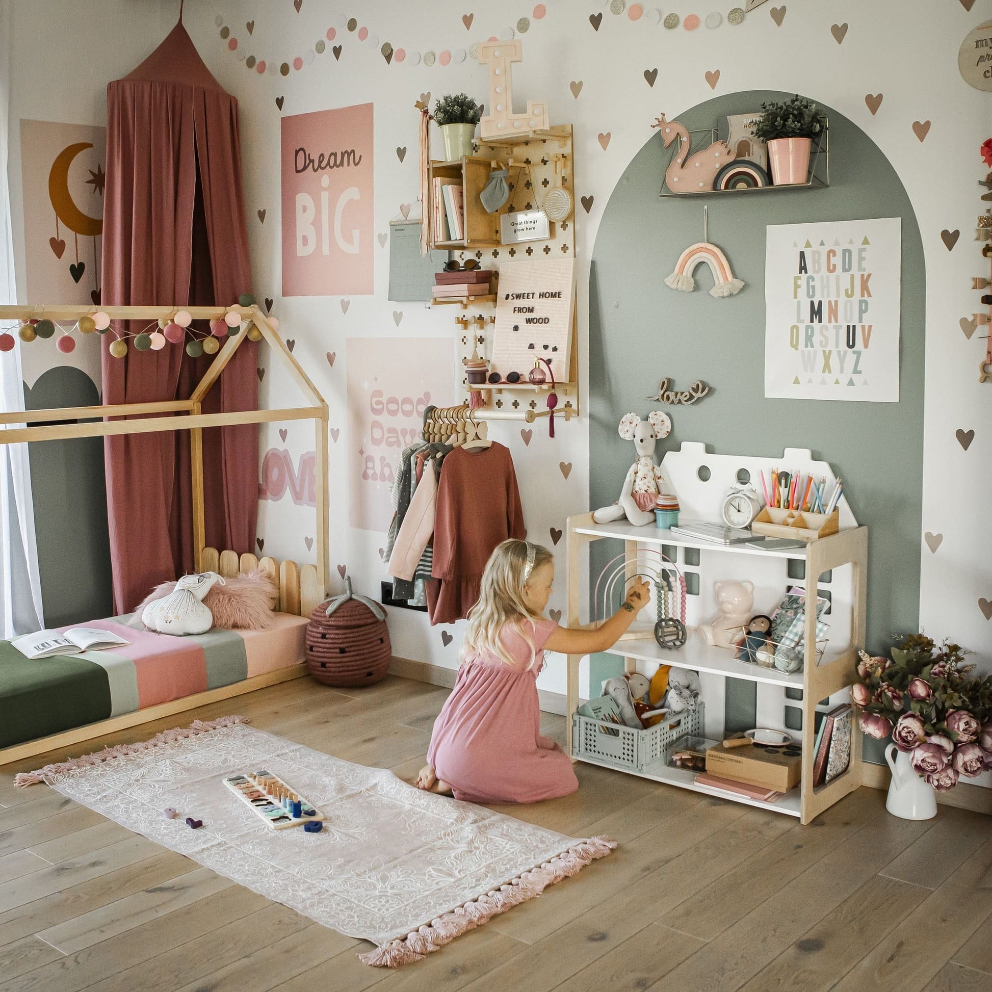 A child in a pink dress sits on the floor of a well-decorated room, playing with a kitchenette set. The space features pastel colors, various toys, and motivational wall art. Nearby, a 2-in-1 doll house and Montessori shelf showcasing an array of toys complete the charming scene.