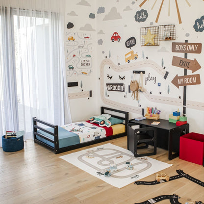 A child's bedroom with a road-themed decor features a toddler floor bed with a horizontal rail headboard and footboard, a black table adorned with toys, and wall decorations featuring cars and signs. A playmat with a road pattern is on the floor, encouraging independent sleeping in this playful space.