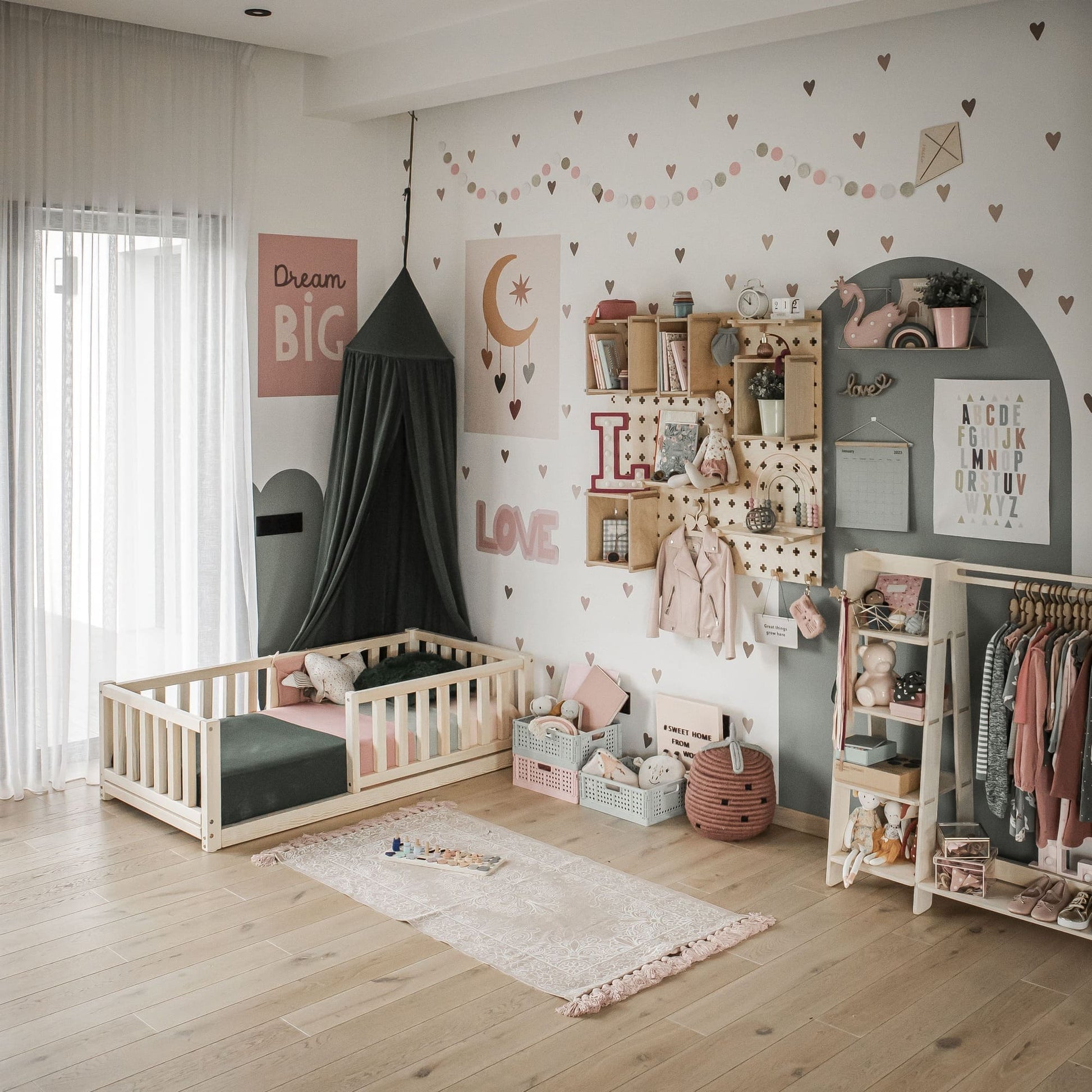 A girl's bedroom featuring a Montessori kids' bed with a fence, wall shelves, a hanging clothes rack, and a canopy. The decor includes heart patterns, a moon and stars, and soft pastel tones. Natural light filters through sheer curtains, creating the perfect setting for comfortable and independent sleeping.