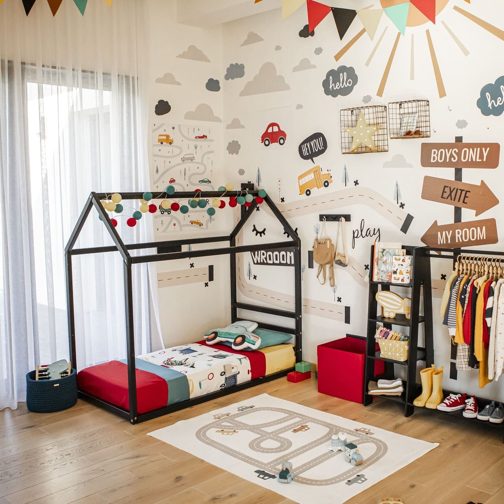 A colorful child's room features a Children's house bed with a horizontal headboard, toys, a clothing rack, a road-themed rug, and various wall decorations including a sun, clouds, cars, and motivational signs.