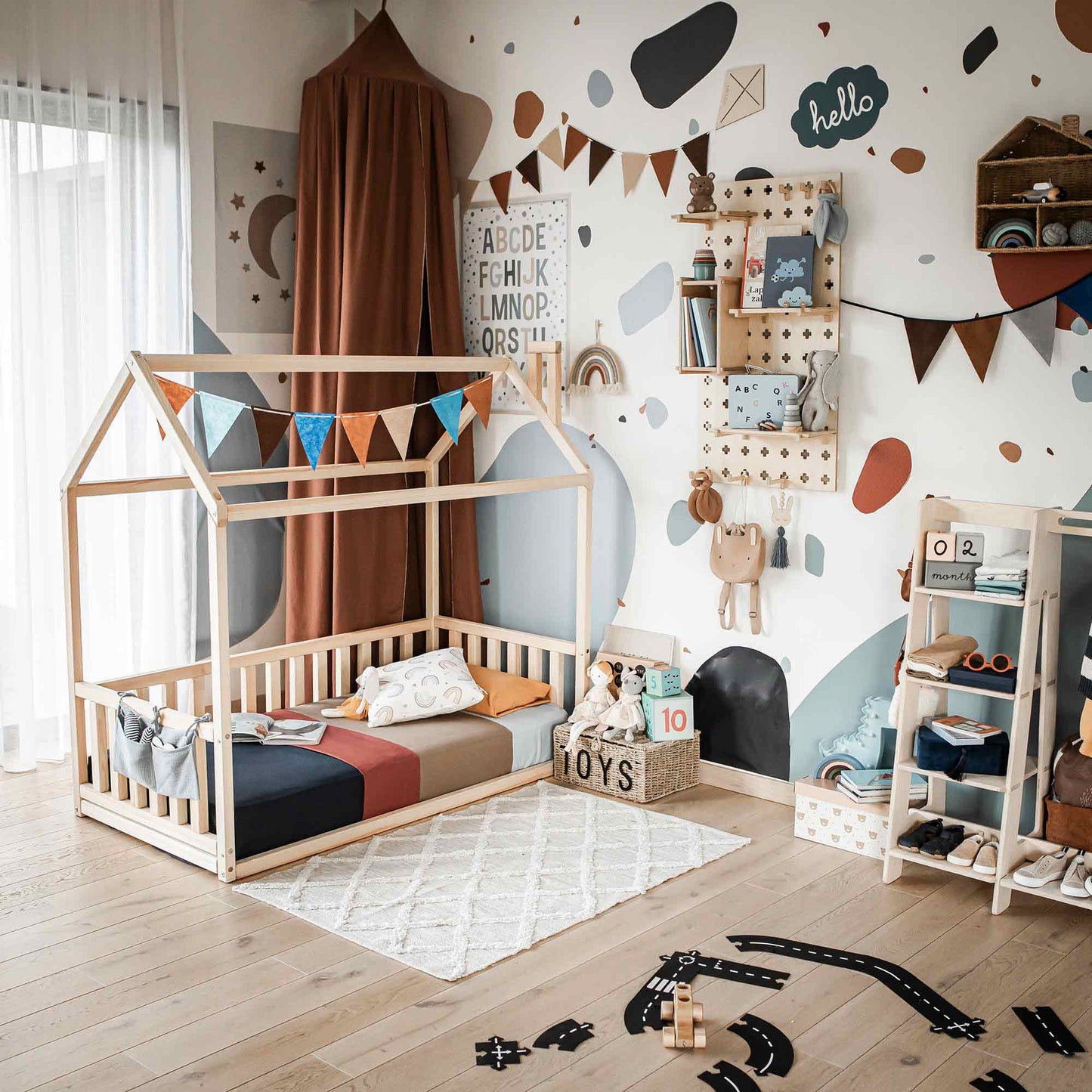A child's bedroom featuring a Kids' house-frame bed with 3-sided rails, bright pillows, a toy chest, shelves, and wall decorations including a banner, alphabet chart, and various art pieces.