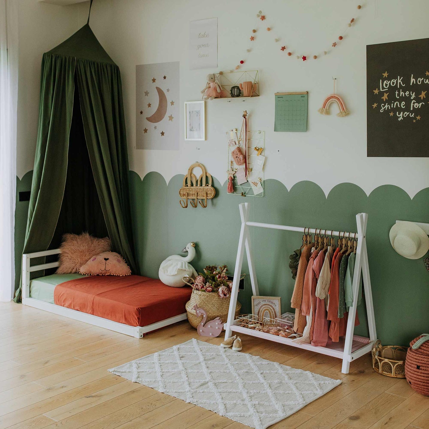 A Montessori bedroom featuring a floor bed, canopy, and wall decorations. The Kids' clothing rack with storage is used as an open Teepee Style Clothing Rack, where children's attire, plush toys, and a woven basket are neatly arranged on a wooden floor with a rug. This setup offers an ideal storage solution for kids.