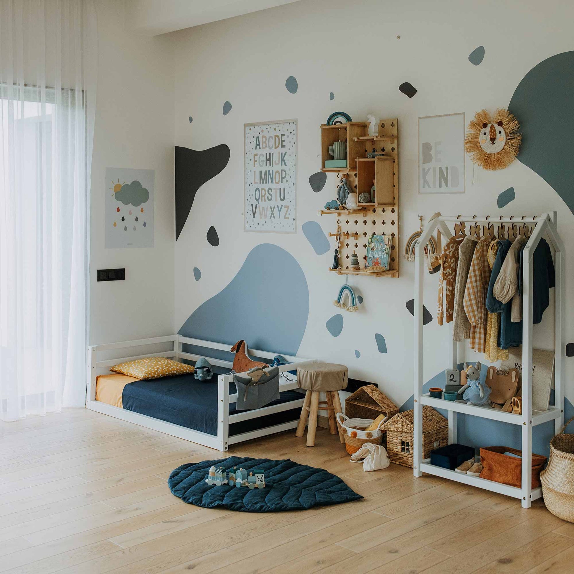 A neatly arranged child's room features a small bed, a Montessori wardrobe adhering to Montessori principles, wall decor with letters and animal designs, and an array of toys and books. White and blue tones dominate the decor.