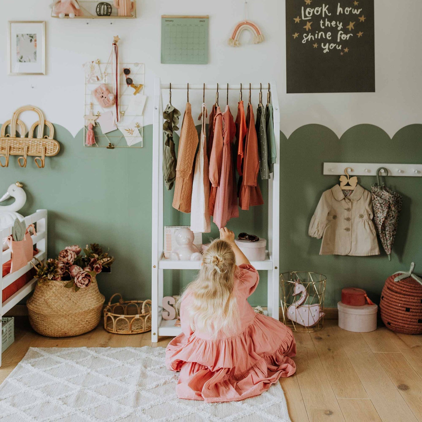 A child in a pink dress is reaching for clothes on a Montessori wardrobe in a neatly decorated room with green and white walls, embodying Montessori principles. The space includes various items like a basket of flowers and a chalkboard.