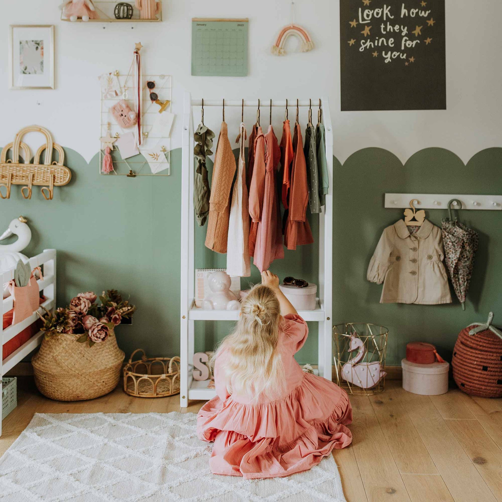 A child in a pink dress is reaching for clothes on a Montessori wardrobe in a neatly decorated room with green and white walls, embodying Montessori principles. The space includes various items like a basket of flowers and a chalkboard.