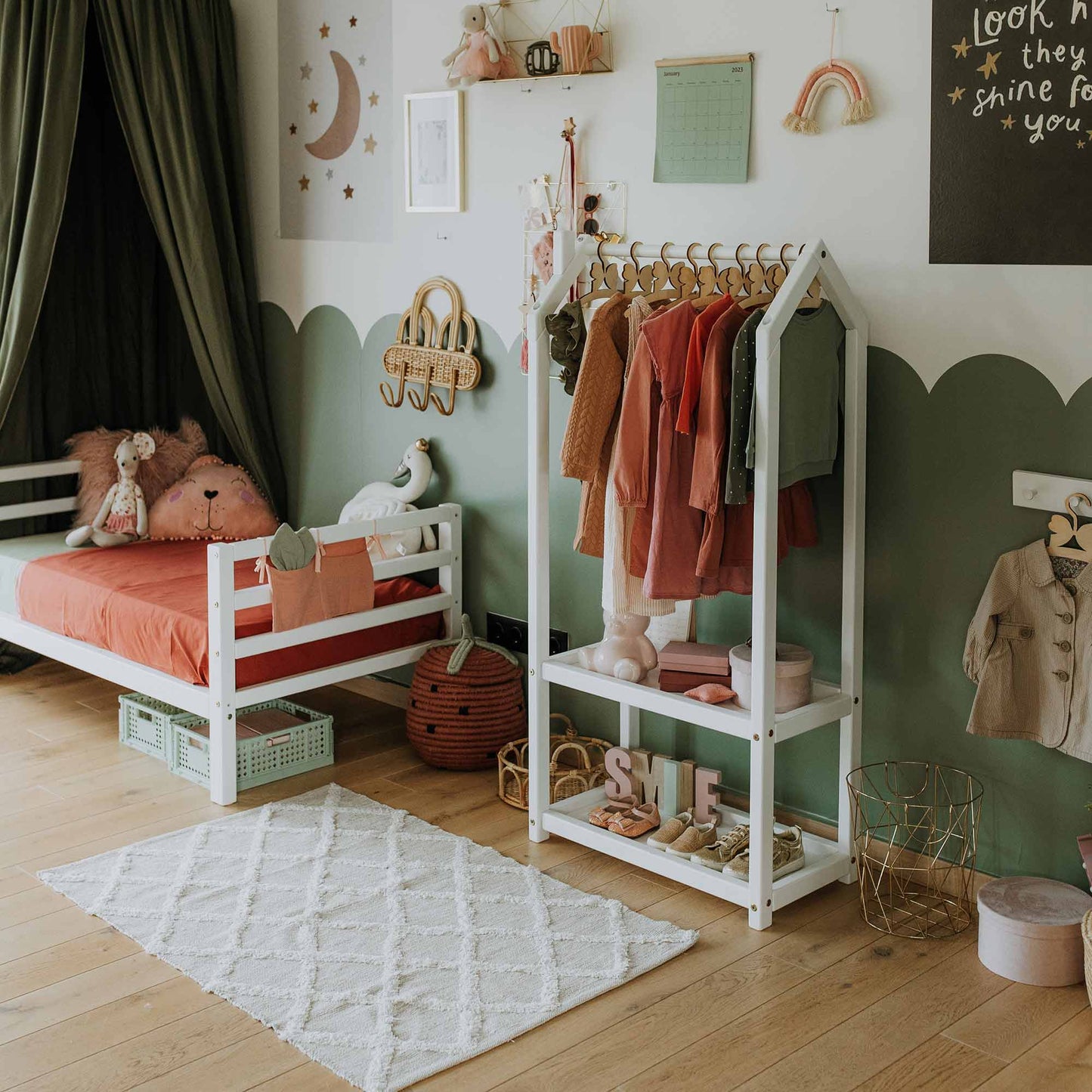 A child's room features a bed, a Montessori wardrobe with hanging clothes, a small shelf labeled "SHINE," an assortment of toys, and wall decorations that include a rainbow and moon. The room has a green and white color scheme.