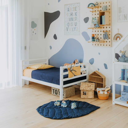 A child's bedroom featuring a kids' bed on legs with a horizontal rail headboard and footboard, wooden shelves, baskets for storage, and educational decorations. The room is designed in a navy blue and light neutral color scheme. The floor is adorned with a blue play mat.