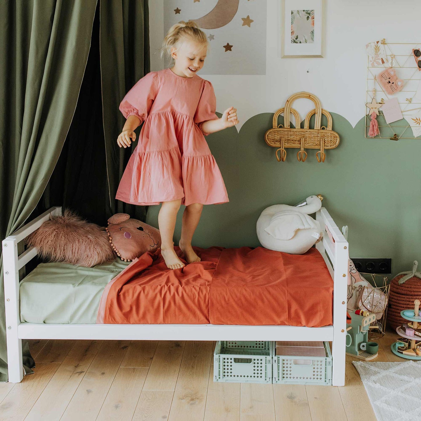 A young child in a pink dress stands on a Kids' bed on legs with a horizontal rail headboard and footboard, in a neatly arranged children's room with green and red bedding, plush toys, and various decorative items on the walls.