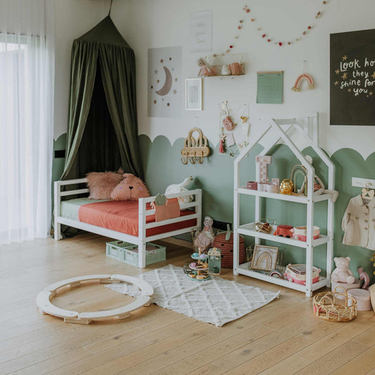 A child's room with a solid wood Montessori toddler bed, featuring the Kids' bed on legs with a horizontal rail headboard and footboard, a green canopy, shelves holding toys and books, a playmat with a toy track, and various decorations. The decor includes star shapes, pastel colors, and a white rug.