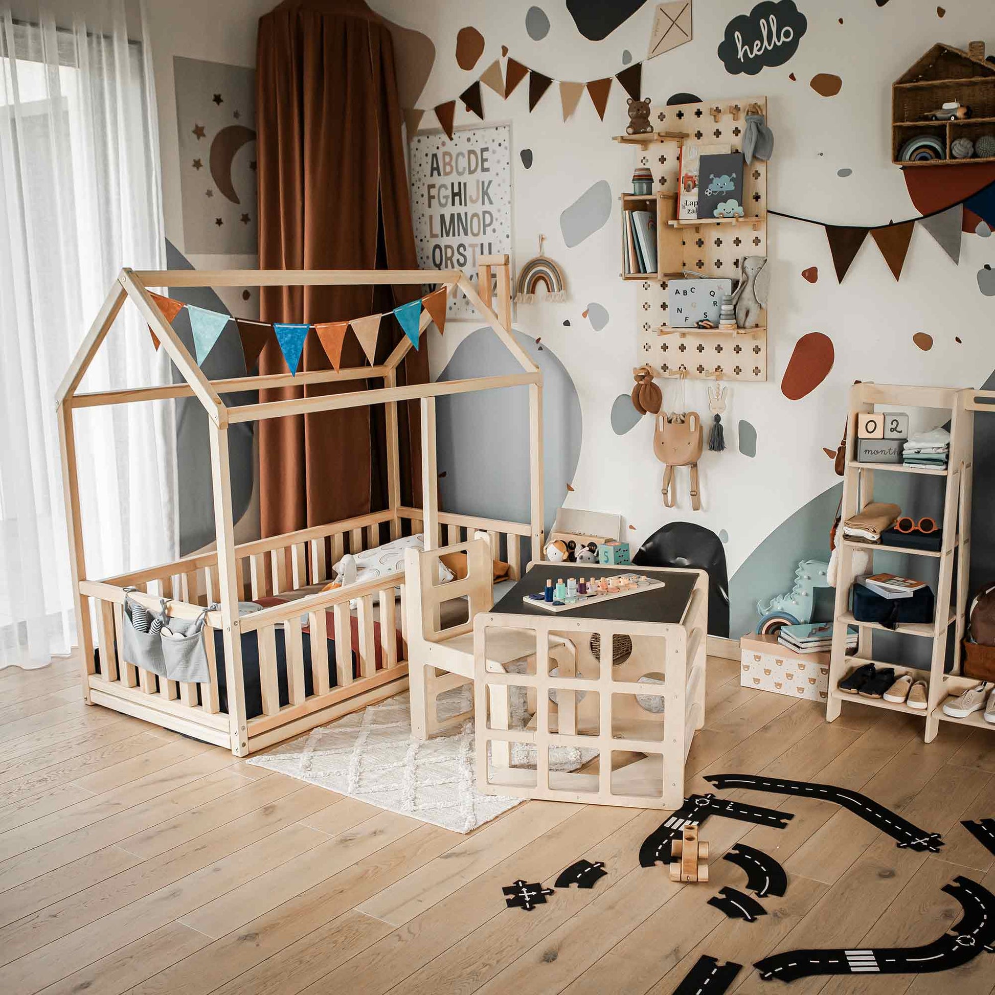 A children's room with a wooden house-shaped bed, a 2-in-1 table and chair set or activity cube in Montessori style, shelves, and colorful decorations. The walls have educational posters and the floor features toys, including a small road mat and a toddler toy for imaginative play.