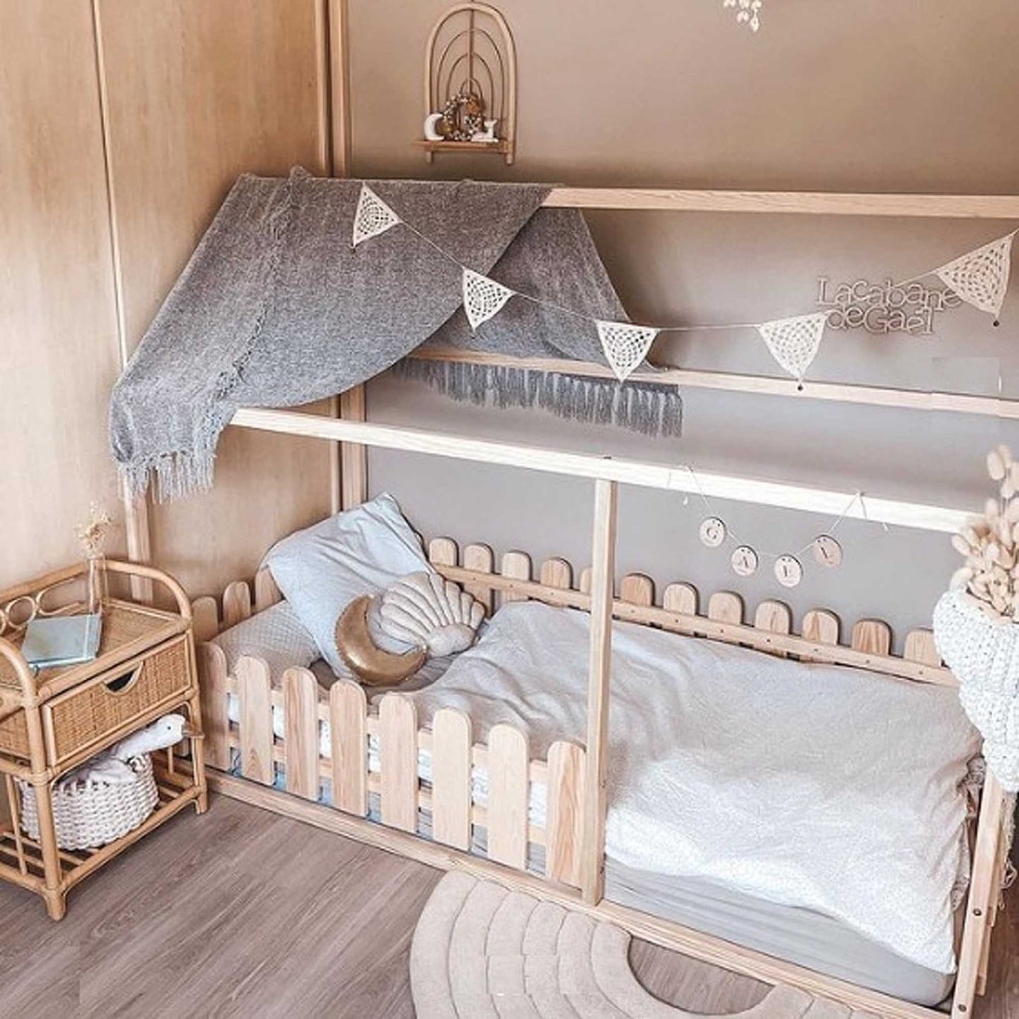 A Platform house bed featuring a picket fence design and a canopy adorned with bunting creates a cozy sleeping area. A wicker nightstand sits beside the toddler bed, holding a toy and some books, in a room that boasts soft, neutral decor.